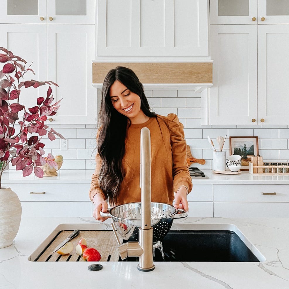 woman holding a colander using a Kraus stainless faucet showing apples, cutting board, knife, utensils, white drawers, dishes